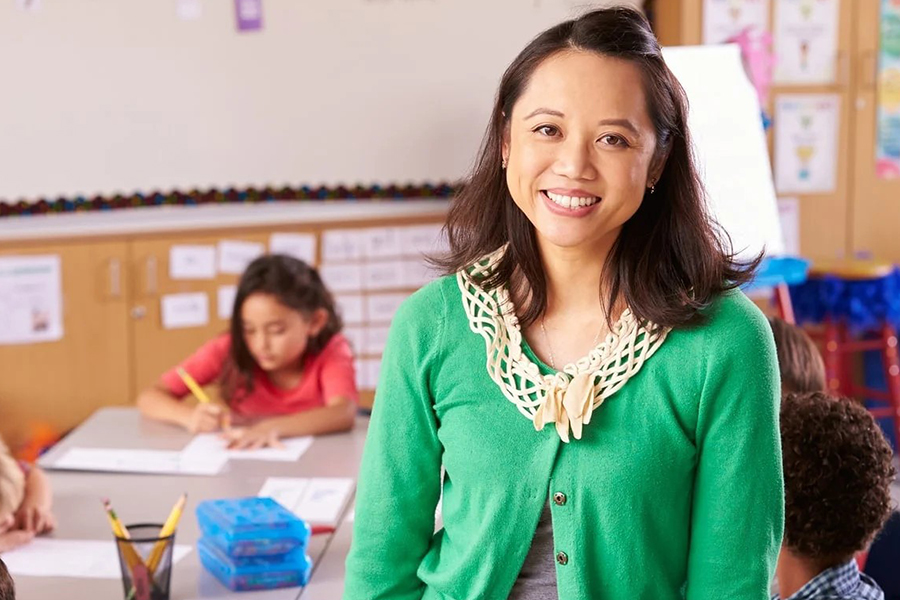 Smiling teacher sits on a desk in an elementary classroom while students write at their desks behind her